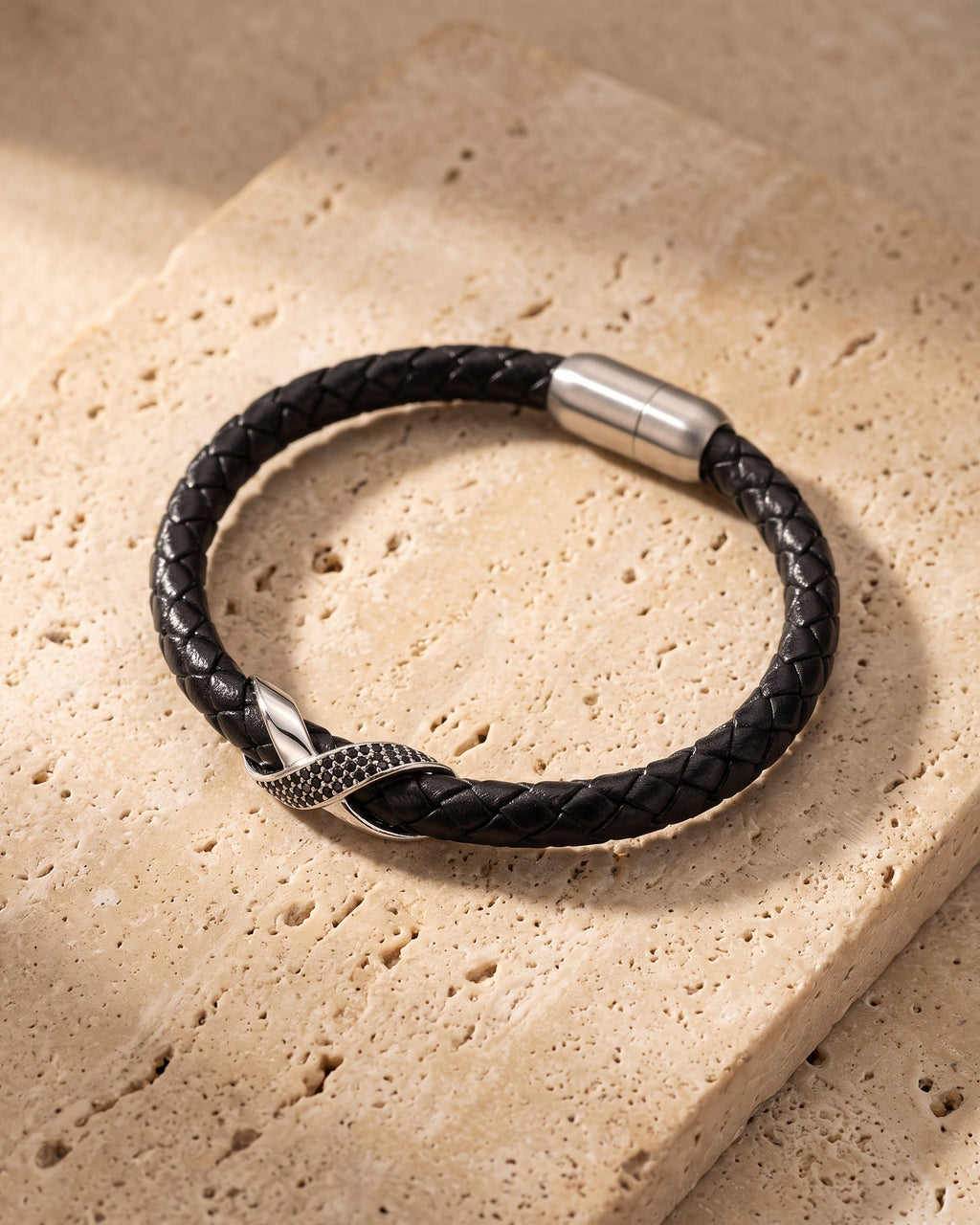 Entire Atlas iced X bracelet in braided leather in silver-tone steel, lying flat on warm stone