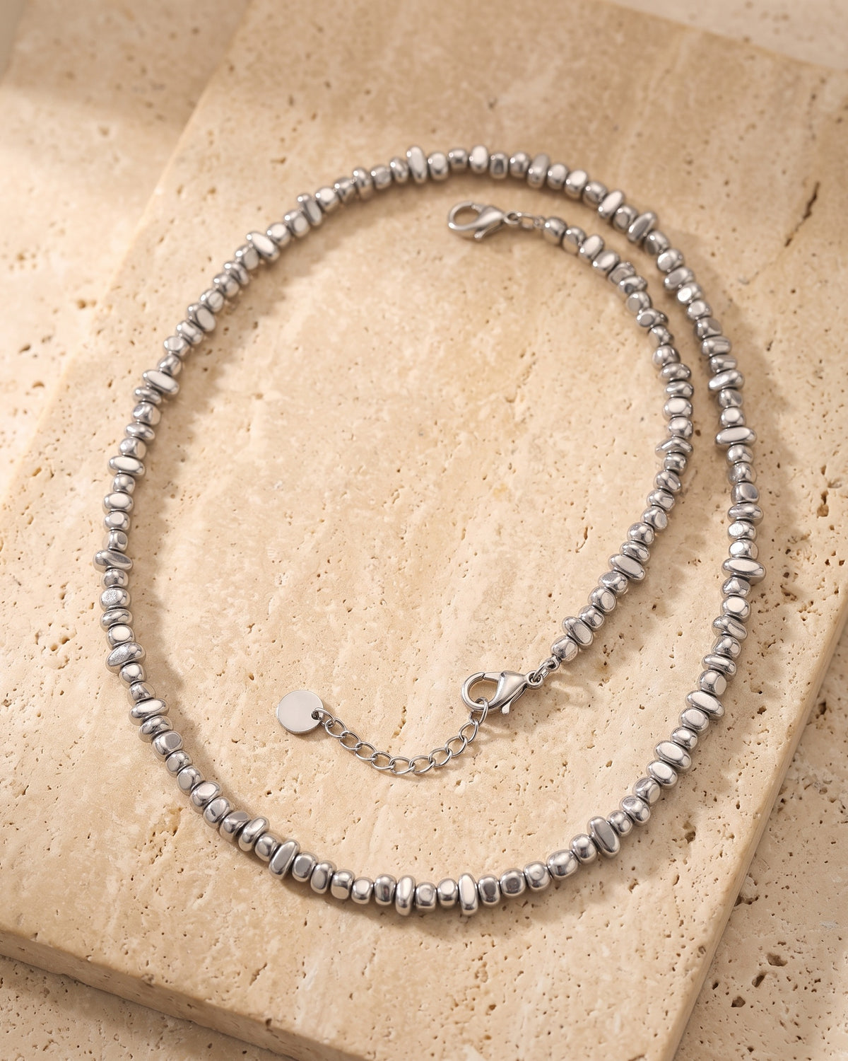 The whole silver Atlas irregular bead chain lying on warm travertine stone