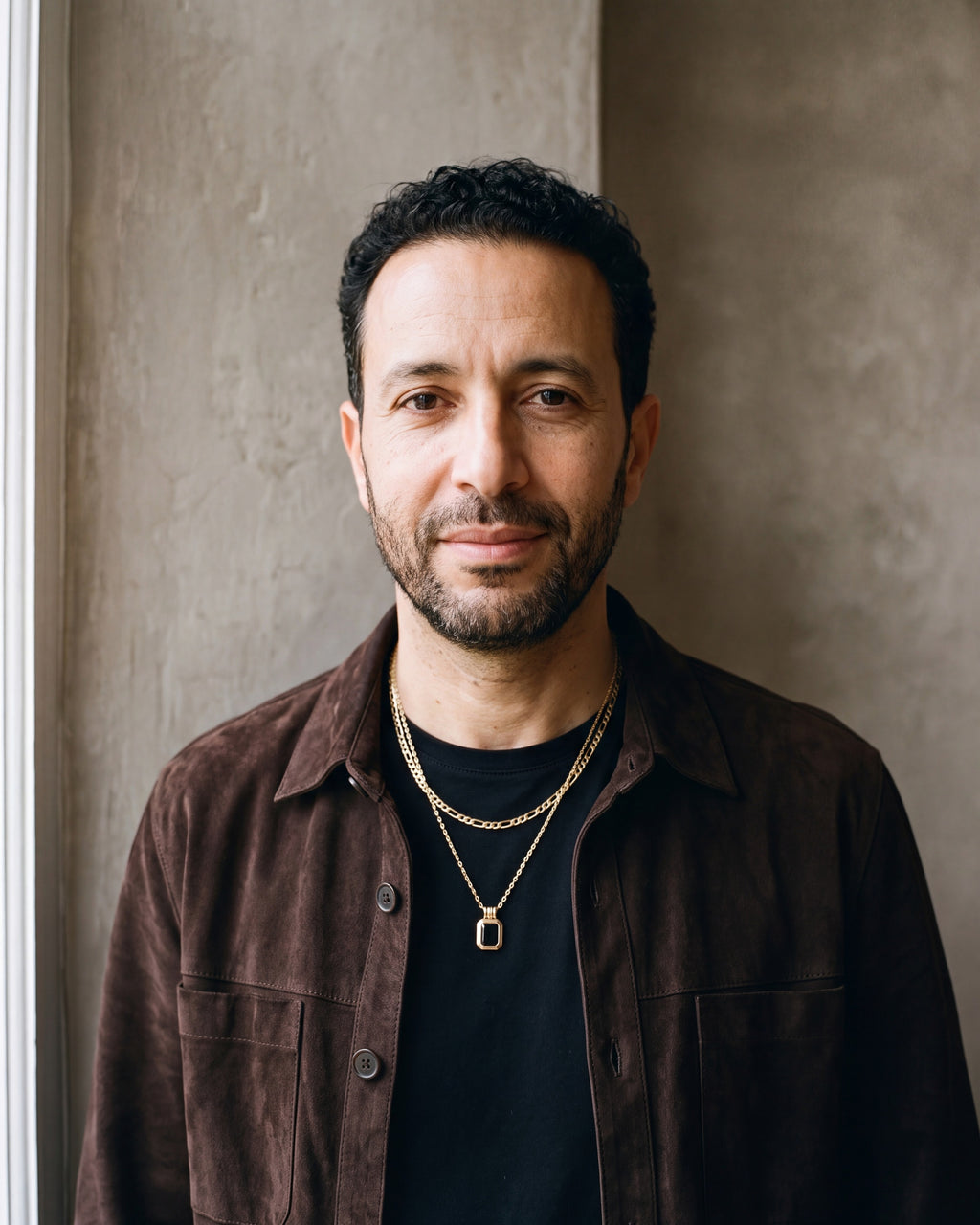 Man in a dark brown suede overshirt open over a black t-shirt by a window wearing the black Atlas Layered Figaro Chain Necklace with Black Stone