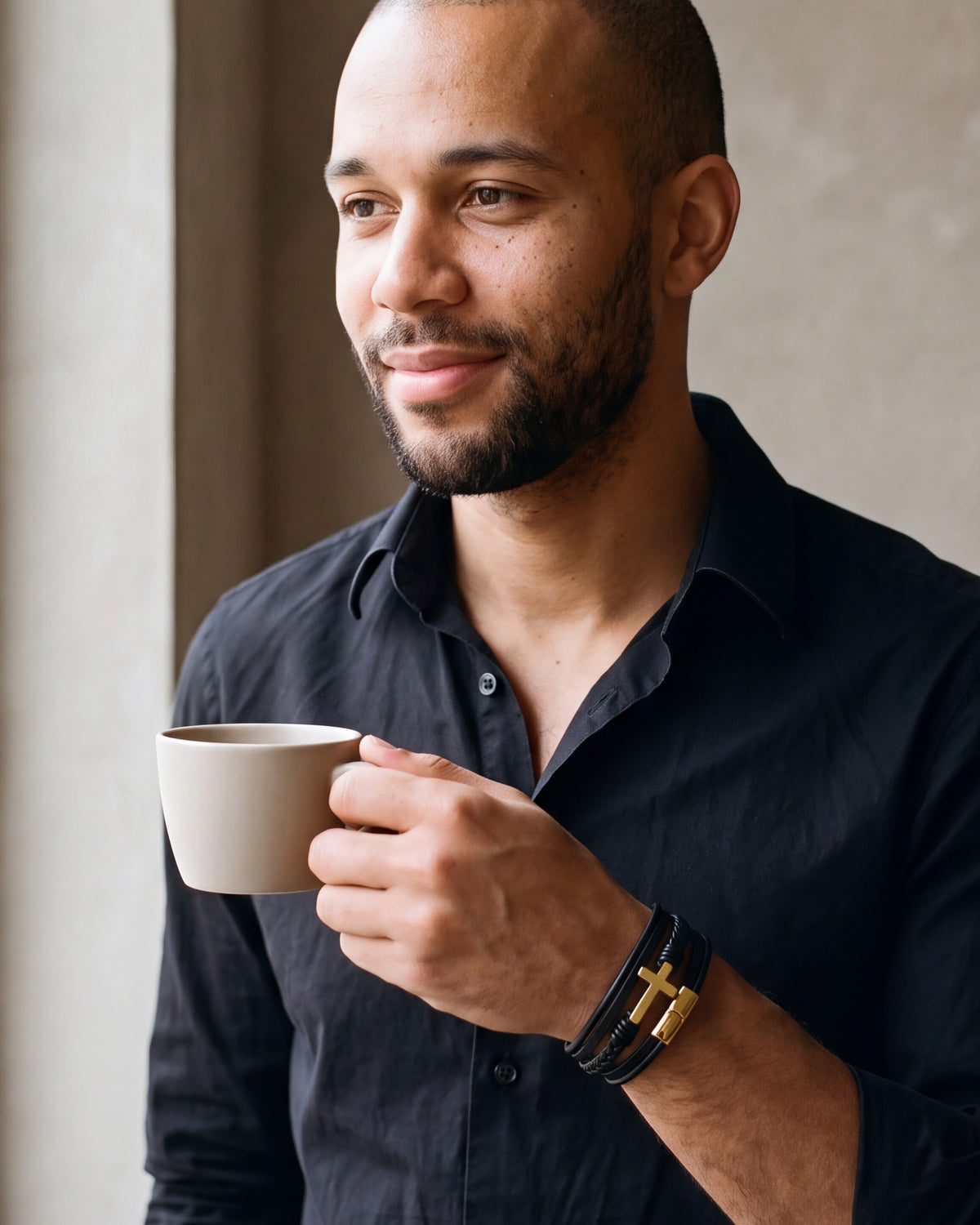 Man in a black shirt holding a cup, wearing the gold-tone Atlas layered leather cross bracelet