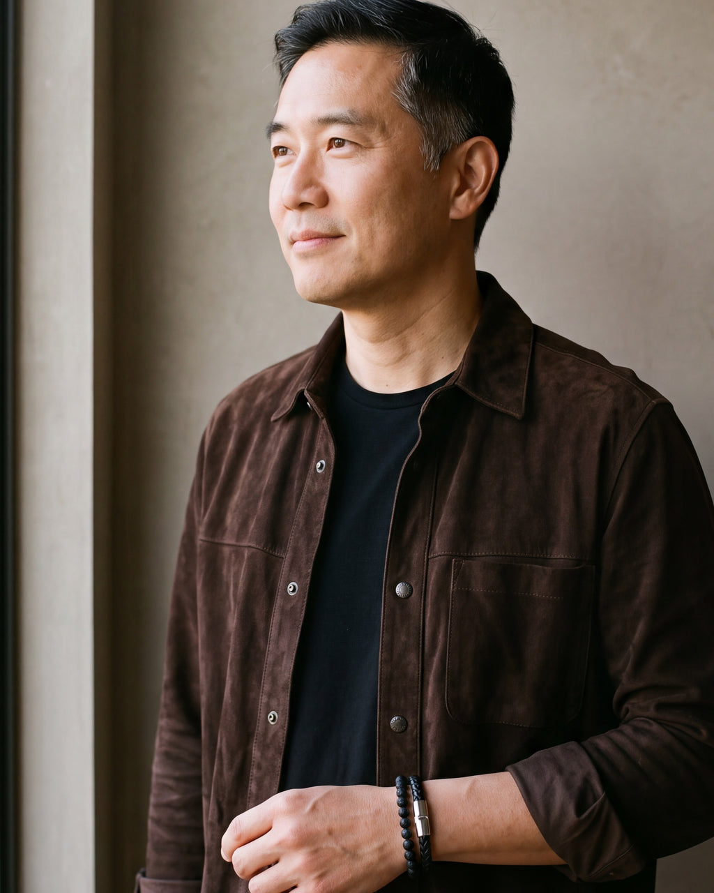 Man in a brown suede overshirt by a window, wearing the Atlas leather and lava stone double bracelet