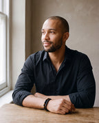 Man in a black shirt seated at a wooden table, wearing the Atlas leather and tiger eye double bracelet