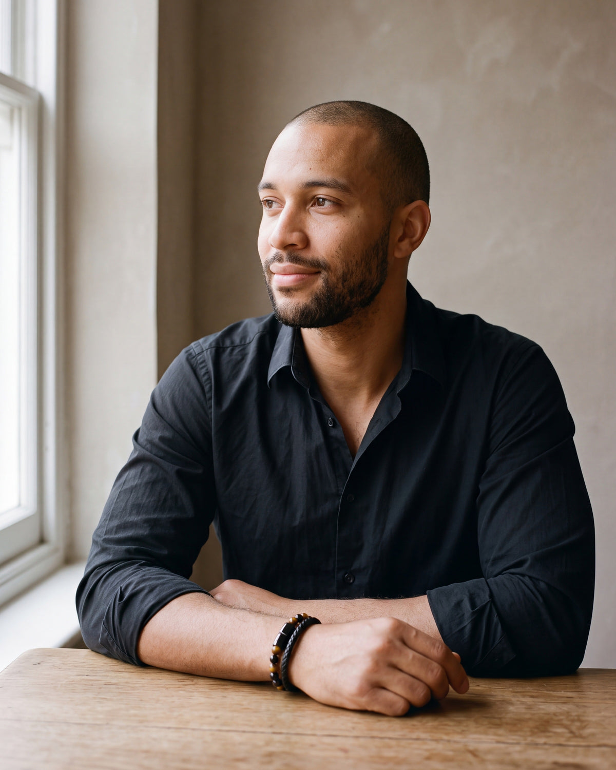 Man in a black shirt seated at a wooden table, wearing the Atlas leather and tiger eye double bracelet