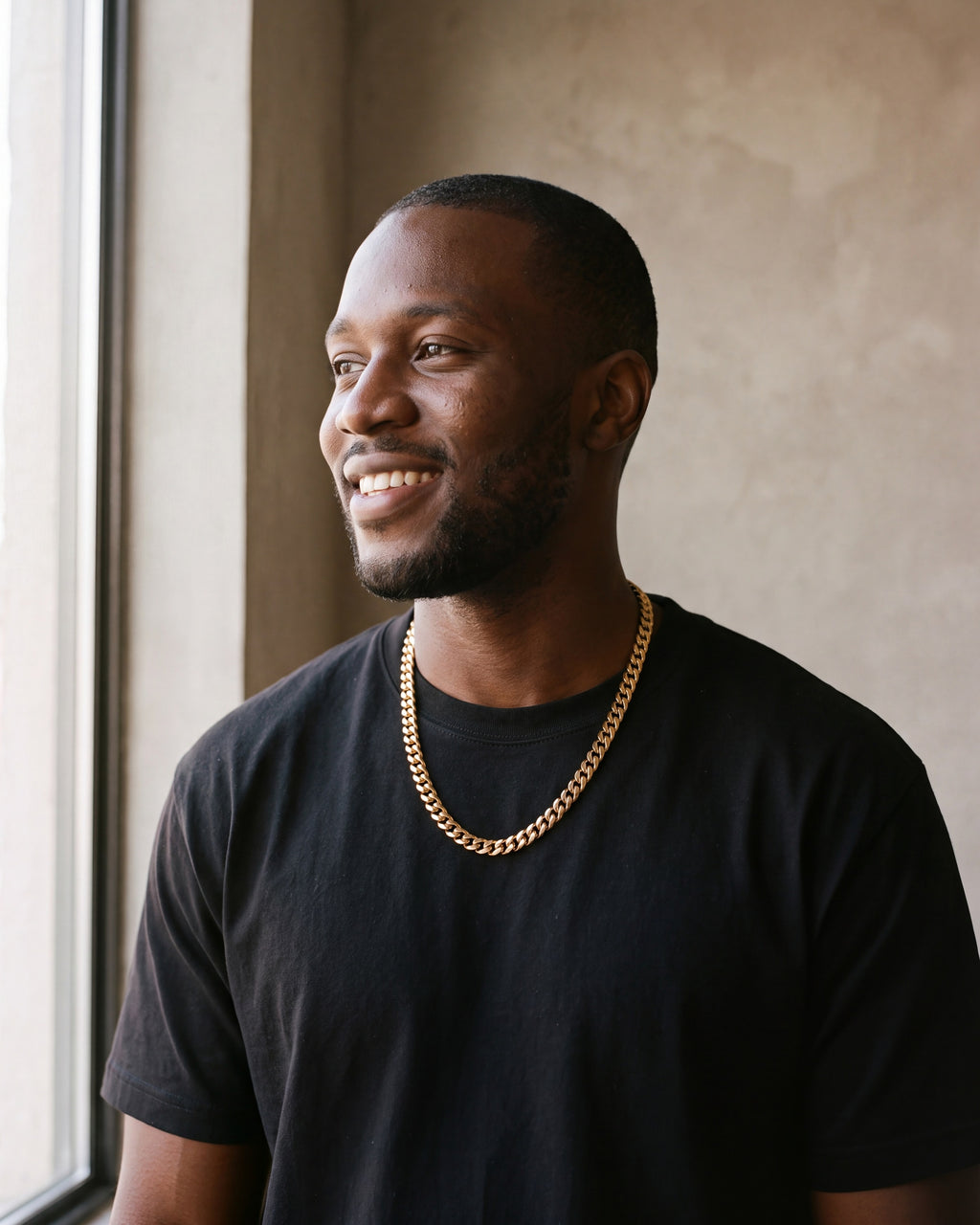 Smiling man by a window wearing the gold Atlas Miami Cuban chain with a black T-shirt