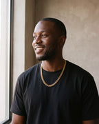 Smiling man by a window wearing the gold Atlas Miami Cuban chain with a black T-shirt