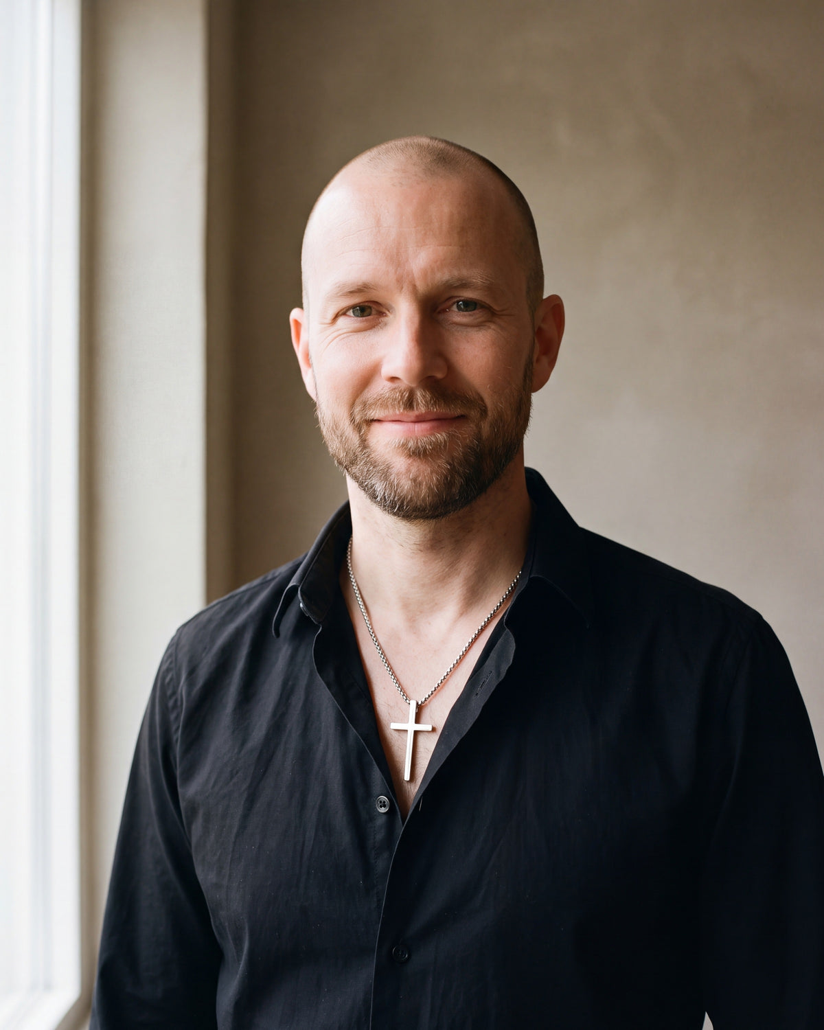 Man in a black shirt by a window wearing the Atlas minimal cross pendant necklace in silver-tone steel