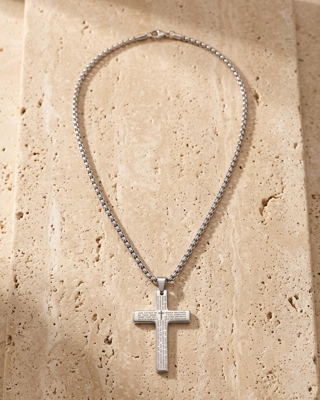 The whole silver-tone Atlas Our Father engraved cross pendant necklace laid on warm stone