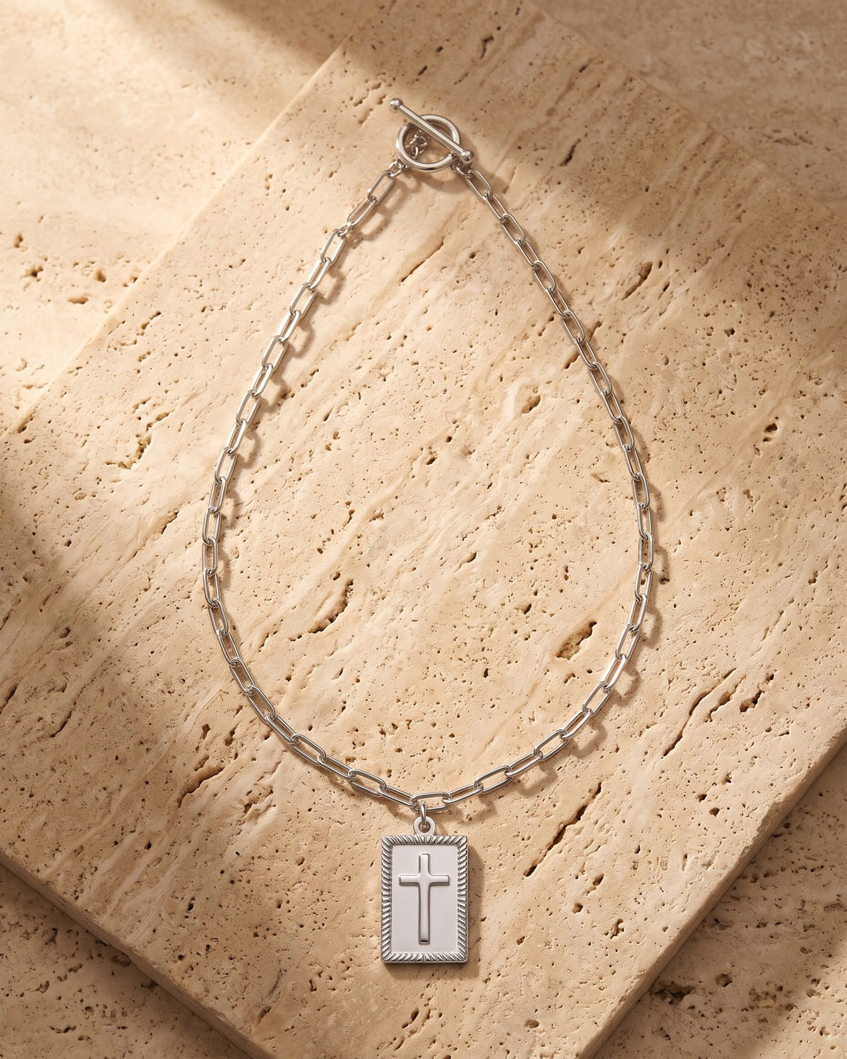 The silver Atlas Paperclip Chain Cross Necklace laid on warm travertine stone