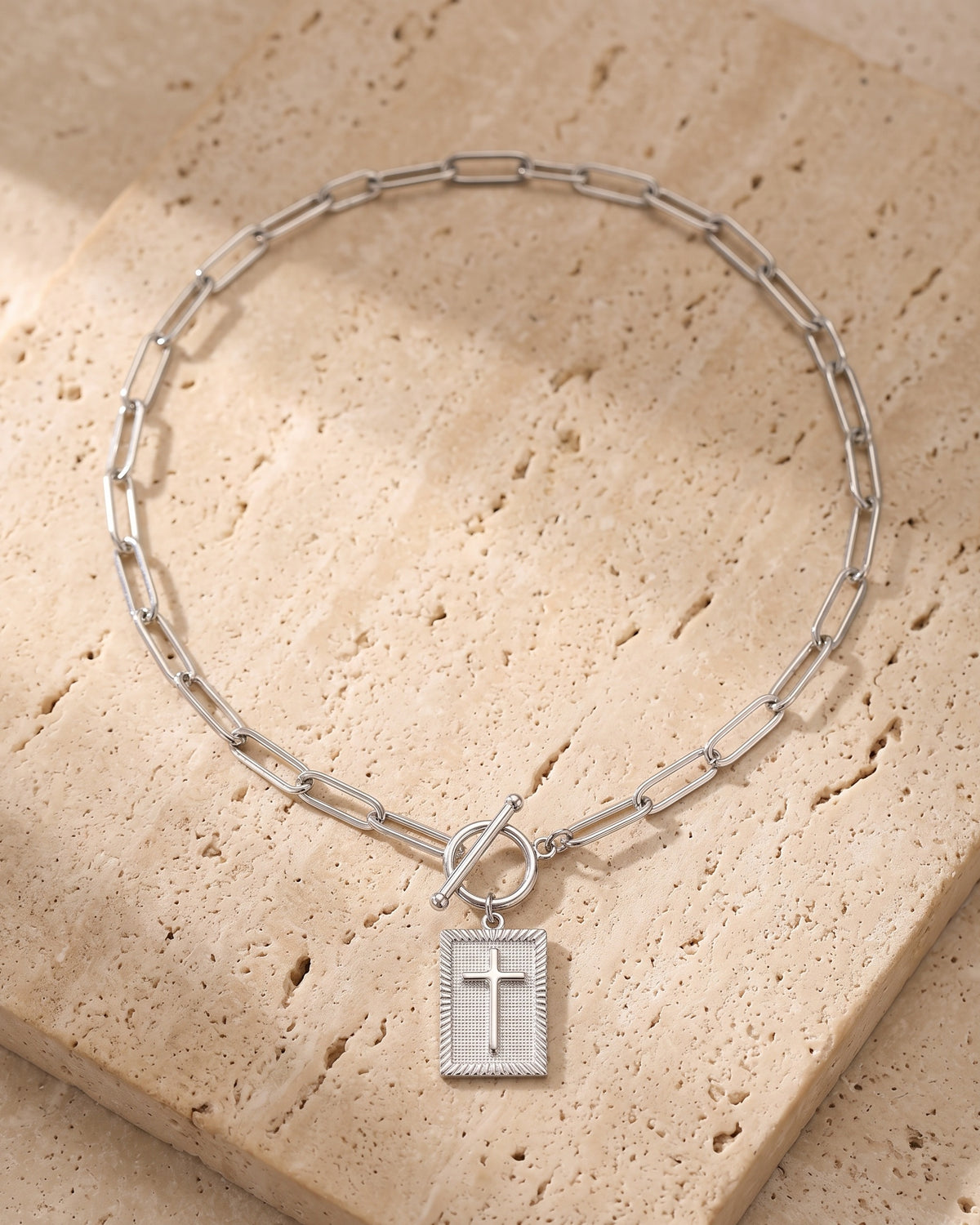 Entire Atlas paperclip chain cross tag necklace in silver textured cross, lying flat on warm stone