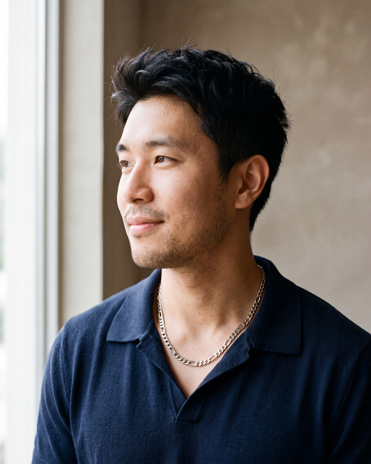 Man in a navy fine-gauge knit polo with an open collar by a window wearing the silver Atlas polished figaro chain