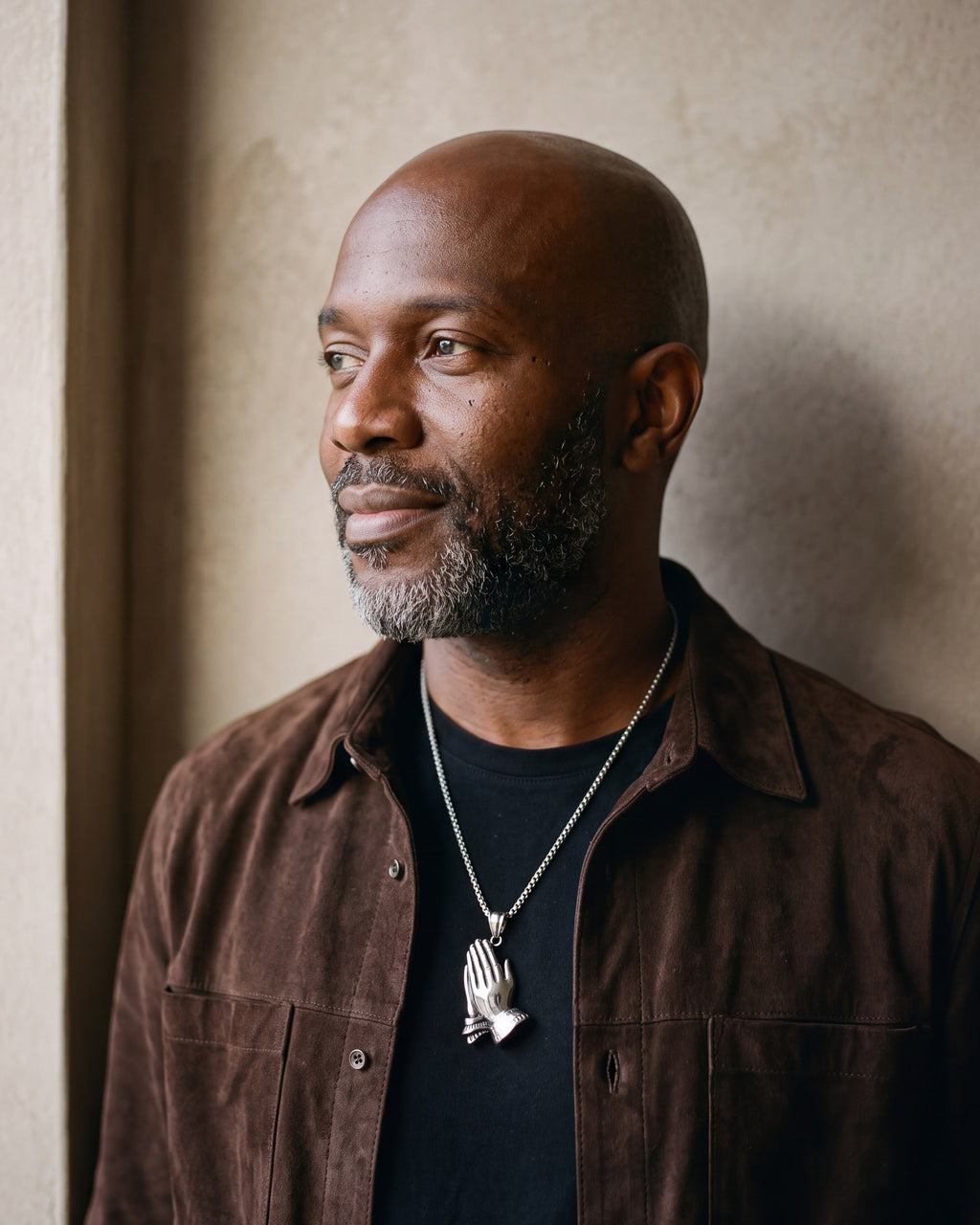 Man in a brown suede overshirt by a window wearing the silver Atlas praying hands pendant necklace