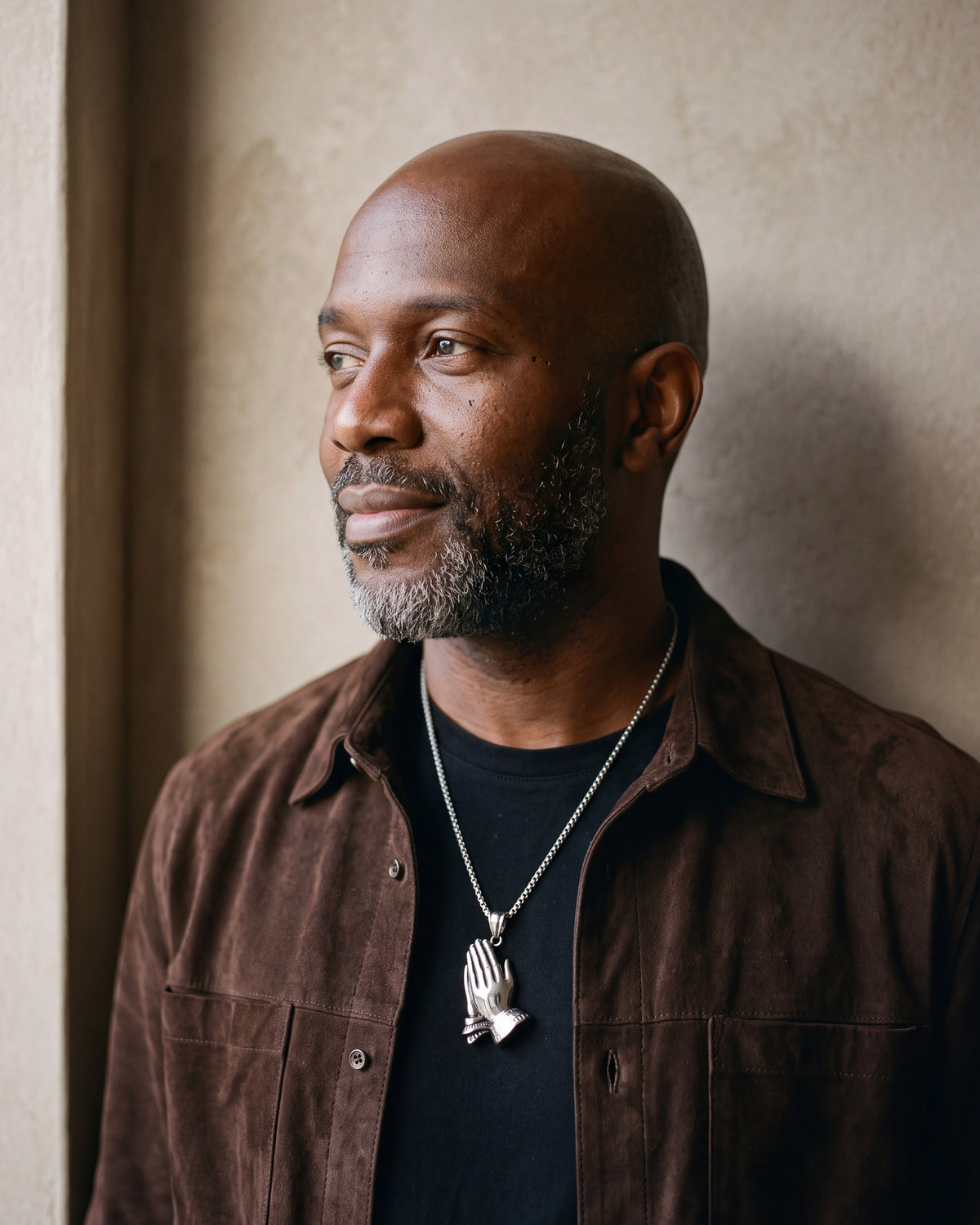 Man in a brown suede overshirt by a window wearing the silver Atlas praying hands pendant necklace