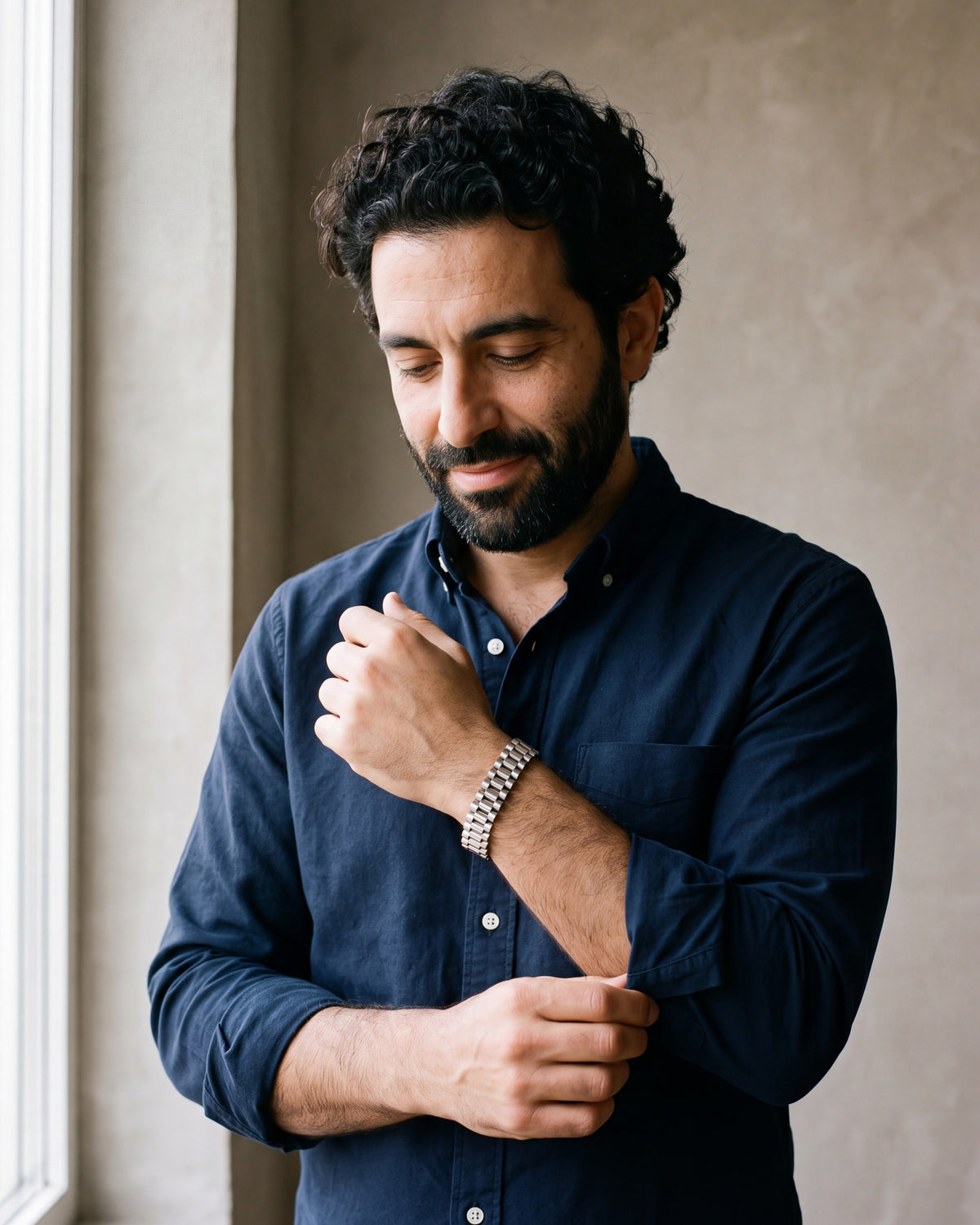 Man in a navy shirt by a window adjusting his cuff, wearing the silver Atlas President link bracelet