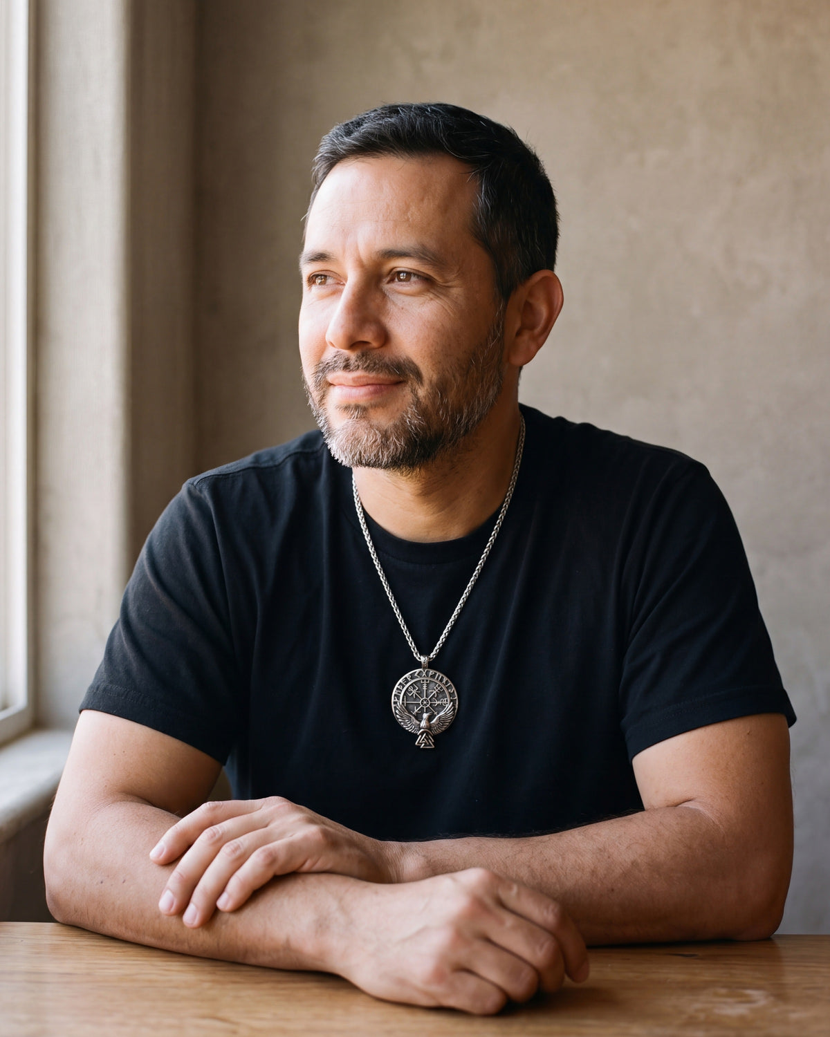 Man in a black T-shirt seated by a window, wearing the Atlas raven Vegvisir compass necklace