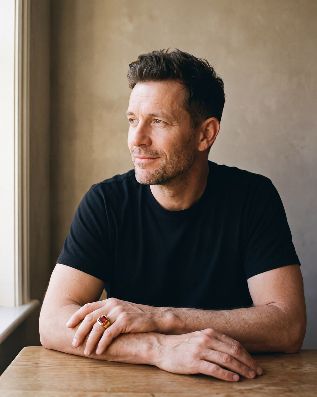 Man in a black T-shirt seated at a wooden table wearing the gold-tone Atlas red stone signet ring