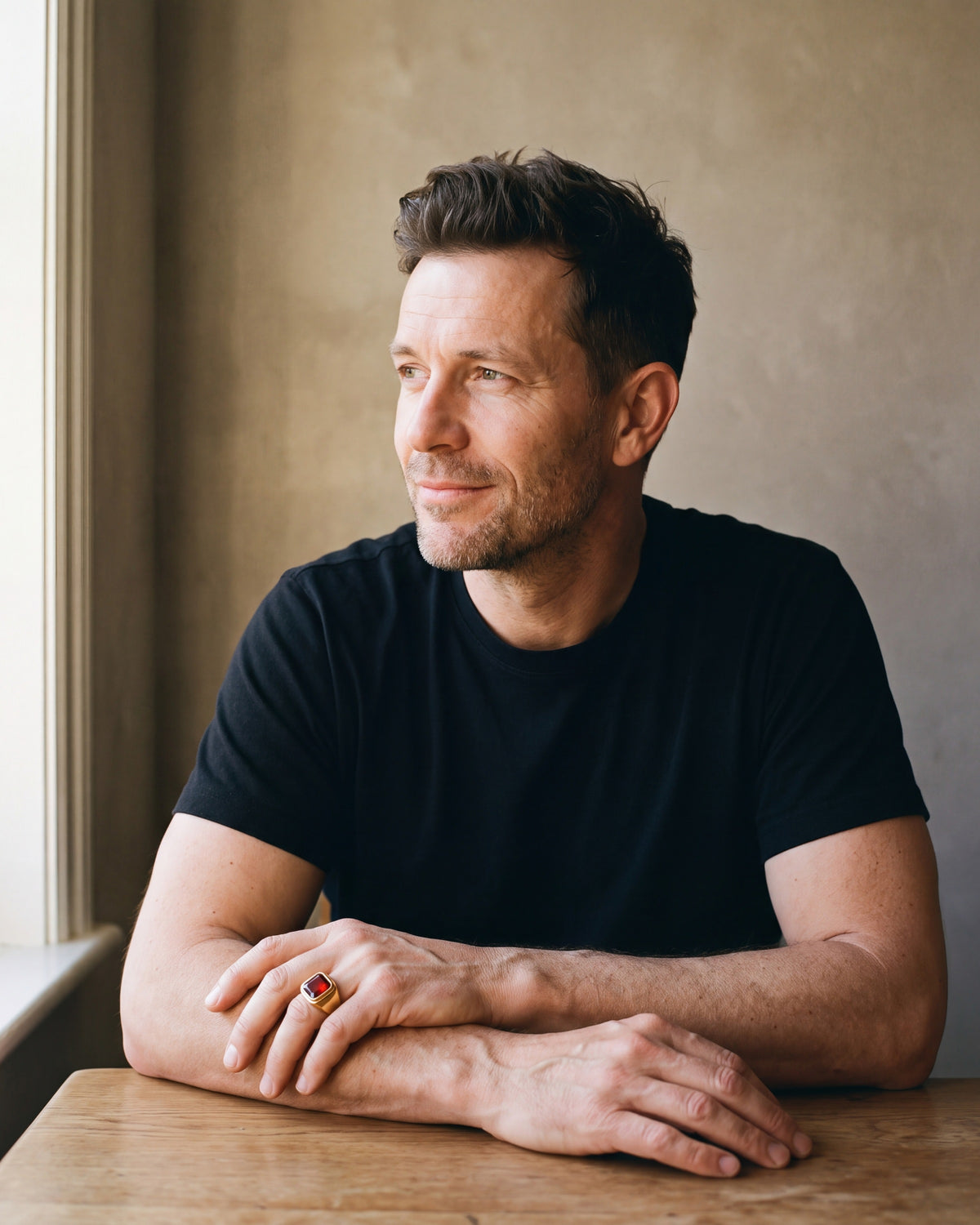 Man in a black T-shirt seated at a wooden table wearing the gold-tone Atlas red stone signet ring