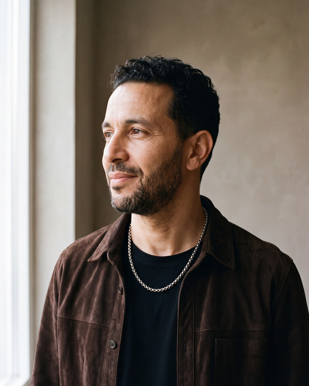 Man in a dark brown suede overshirt open over a black t-shirt by a window wearing the silver-tone Atlas Rolo Chain