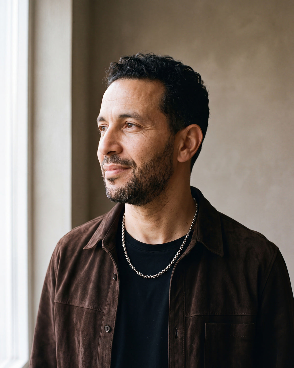 Man in a dark brown suede overshirt open over a black t-shirt by a window wearing the silver-tone Atlas Rolo Chain