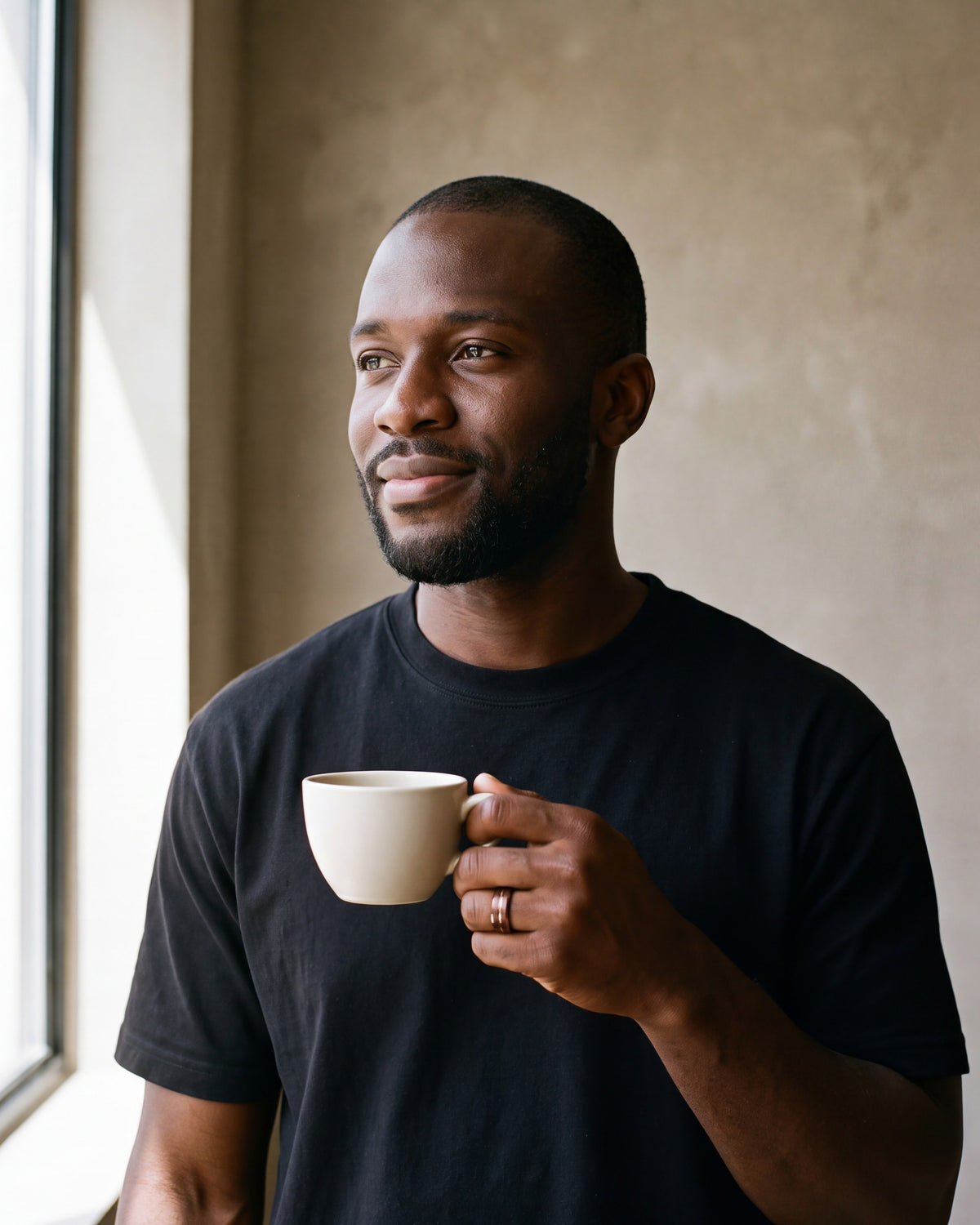Man in a heavyweight black cotton crew-neck t-shirt by a window wearing the rose gold-tone Atlas Rose Gold-Tone Groove Ring