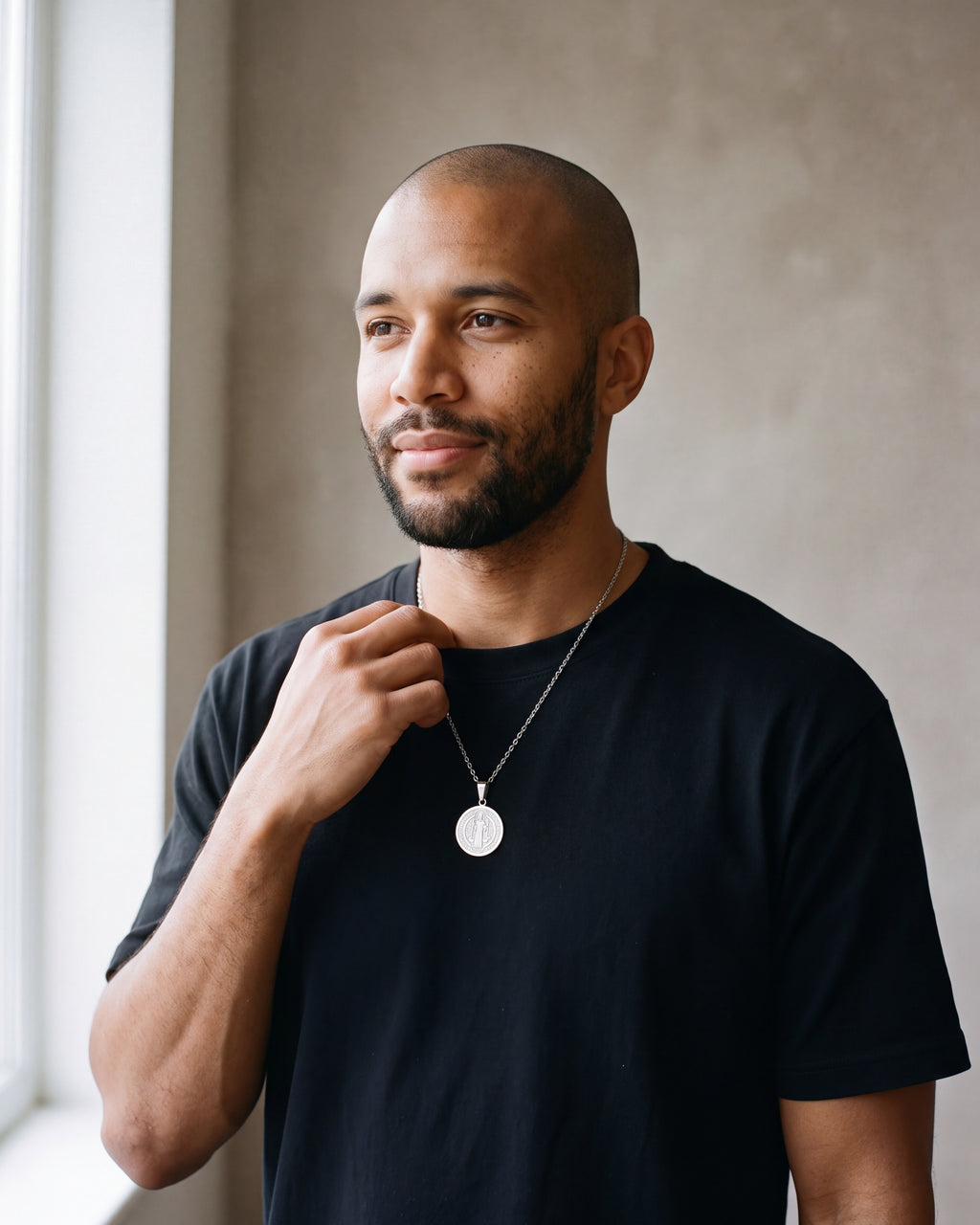 Man in a black merino sweater by a window wearing the Atlas two-sided Saint Benedict medal necklace in silver-tone steel