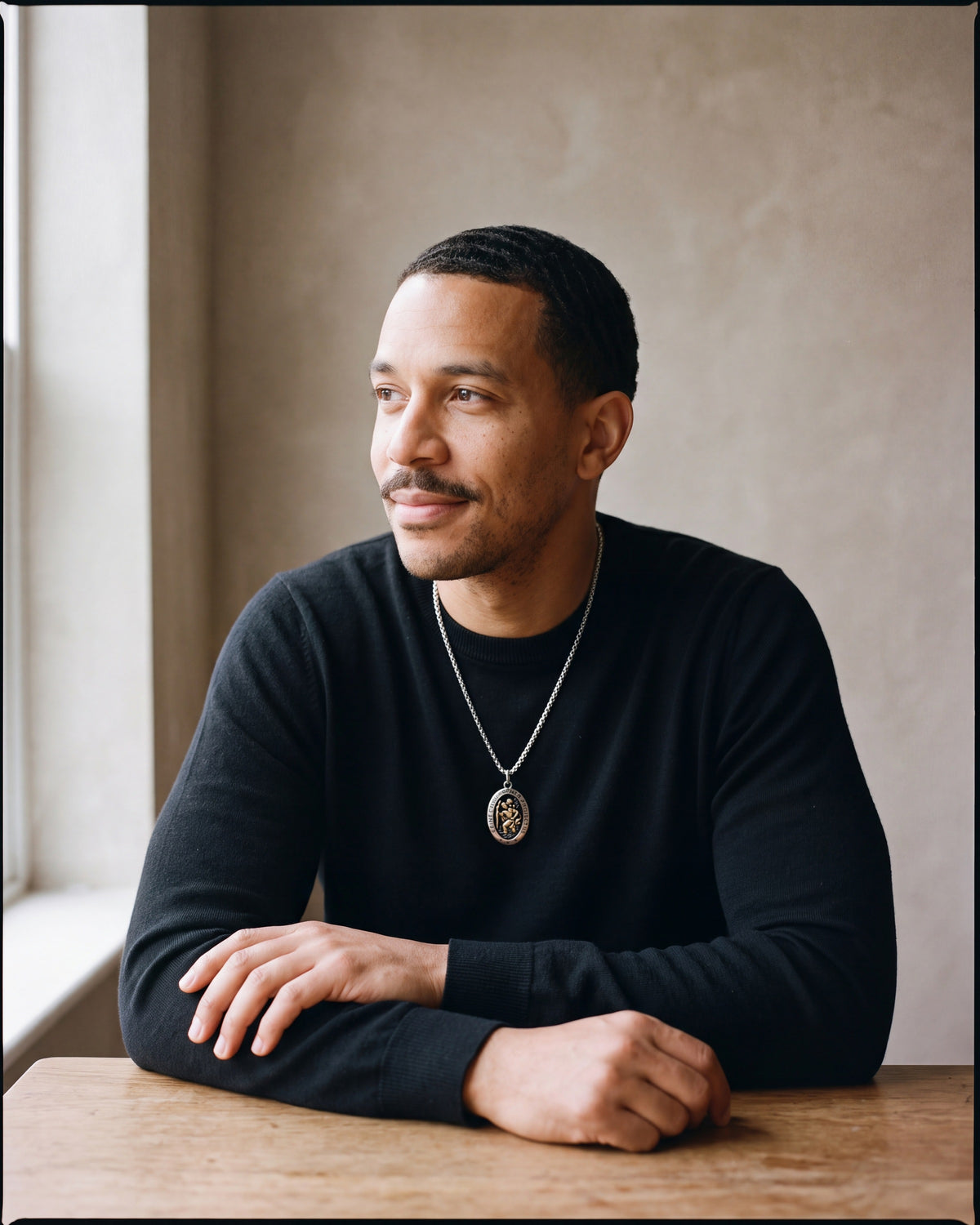 Man in a black merino sweater seated at a wooden table wearing the silver-tone Atlas Saint Christopher oval pendant necklace