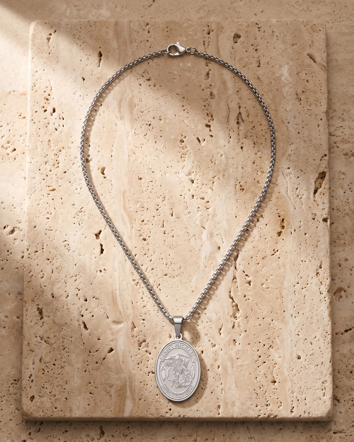 The silver Atlas Saint Michael Pendant Necklace laid on warm travertine stone