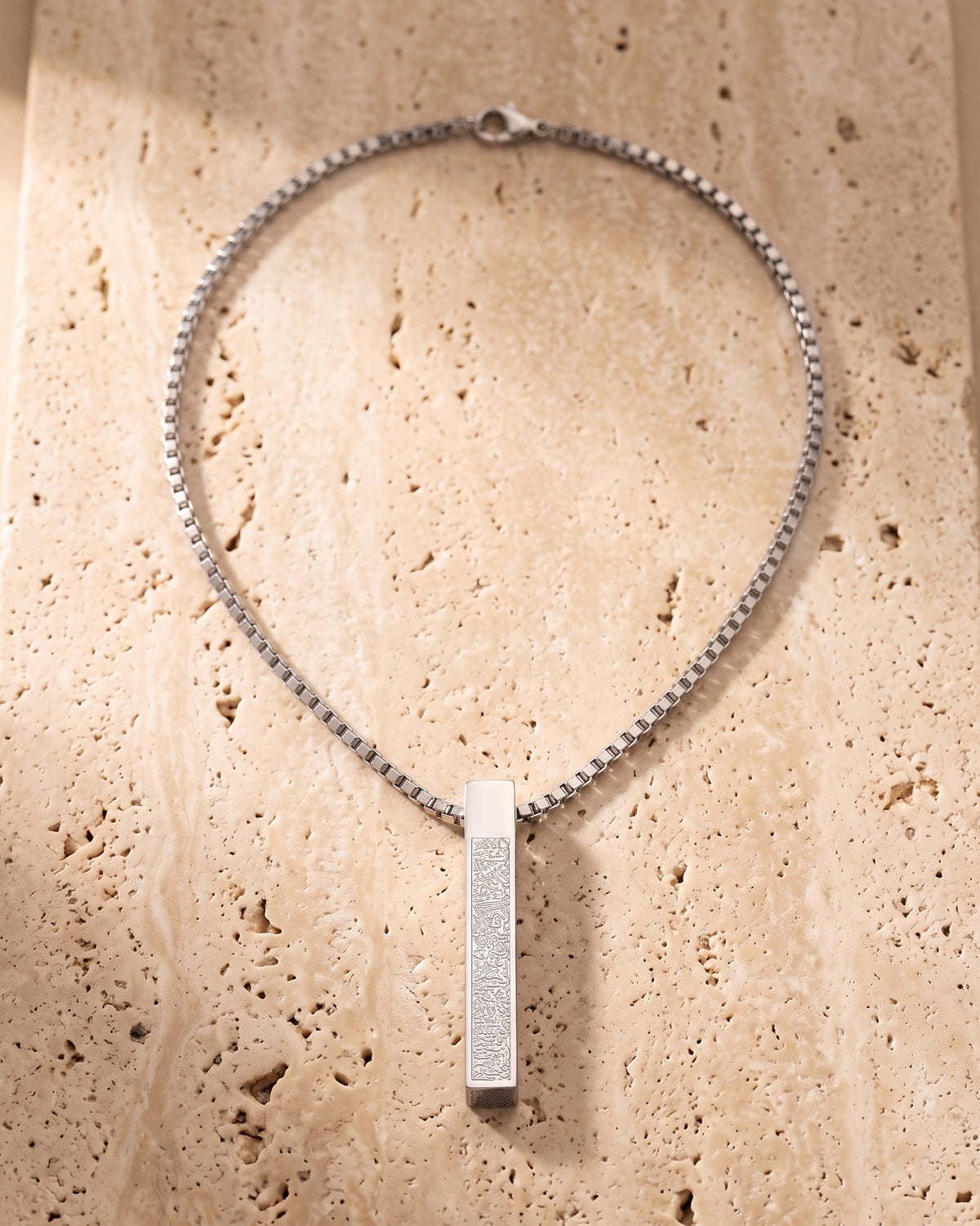 The whole silver-tone Atlas script bar pendant necklace lying on warm travertine stone