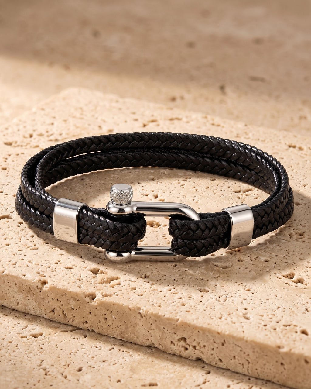 The whole silver-tone / black Atlas Shackle Bracelet in Braided Leather lying on warm travertine stone