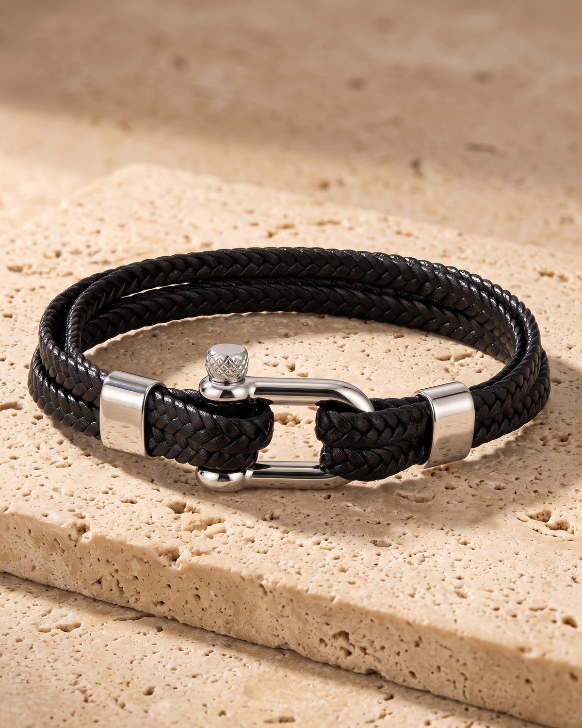The whole silver-tone / black Atlas Shackle Bracelet in Braided Leather lying on warm travertine stone