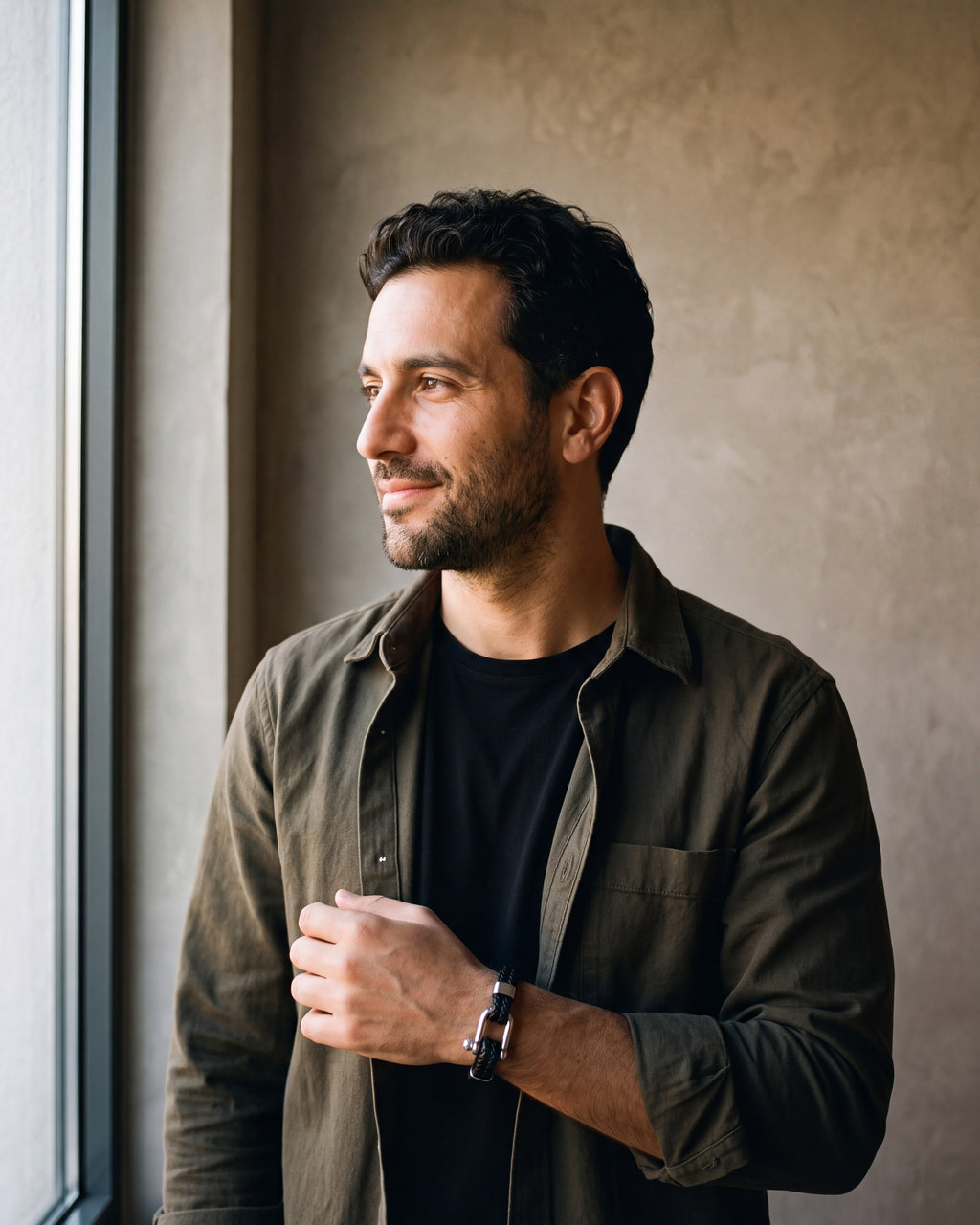 Man in a dark olive overshirt open over a black t-shirt by a window wearing the silver-tone / black Atlas Shackle Bracelet in Braided Leather
