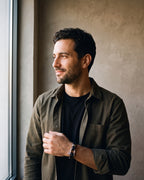 Man in a dark olive overshirt open over a black t-shirt by a window wearing the silver-tone / black Atlas Shackle Bracelet in Braided Leather