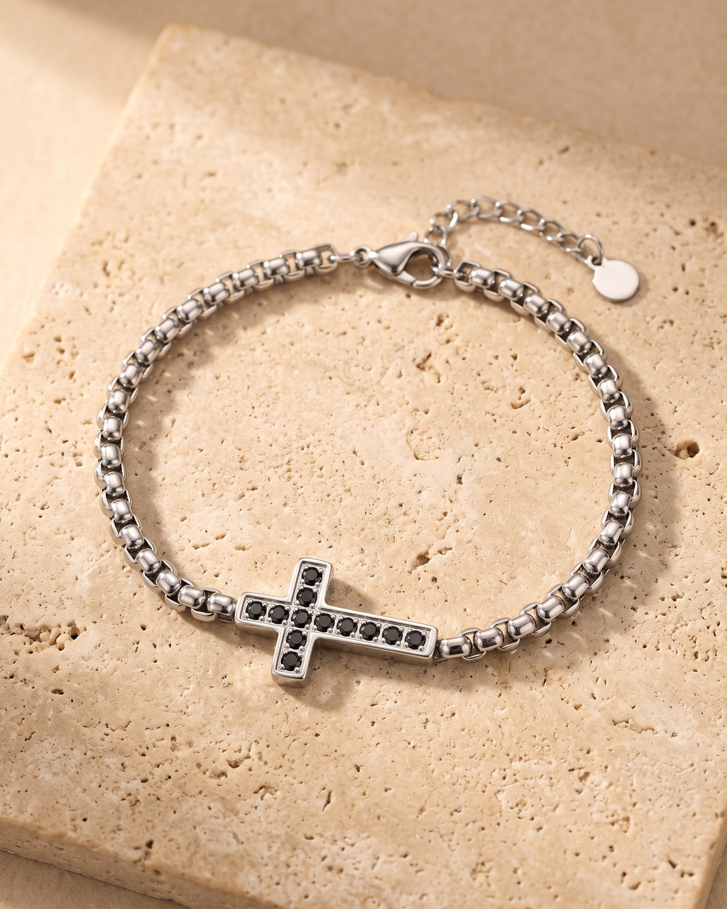 Entire Atlas sideways cross bracelet in silver-tone steel, lying flat on warm stone