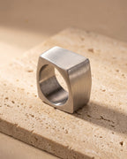 Entire Atlas square top ring in silver-tone steel, lying flat on warm stone