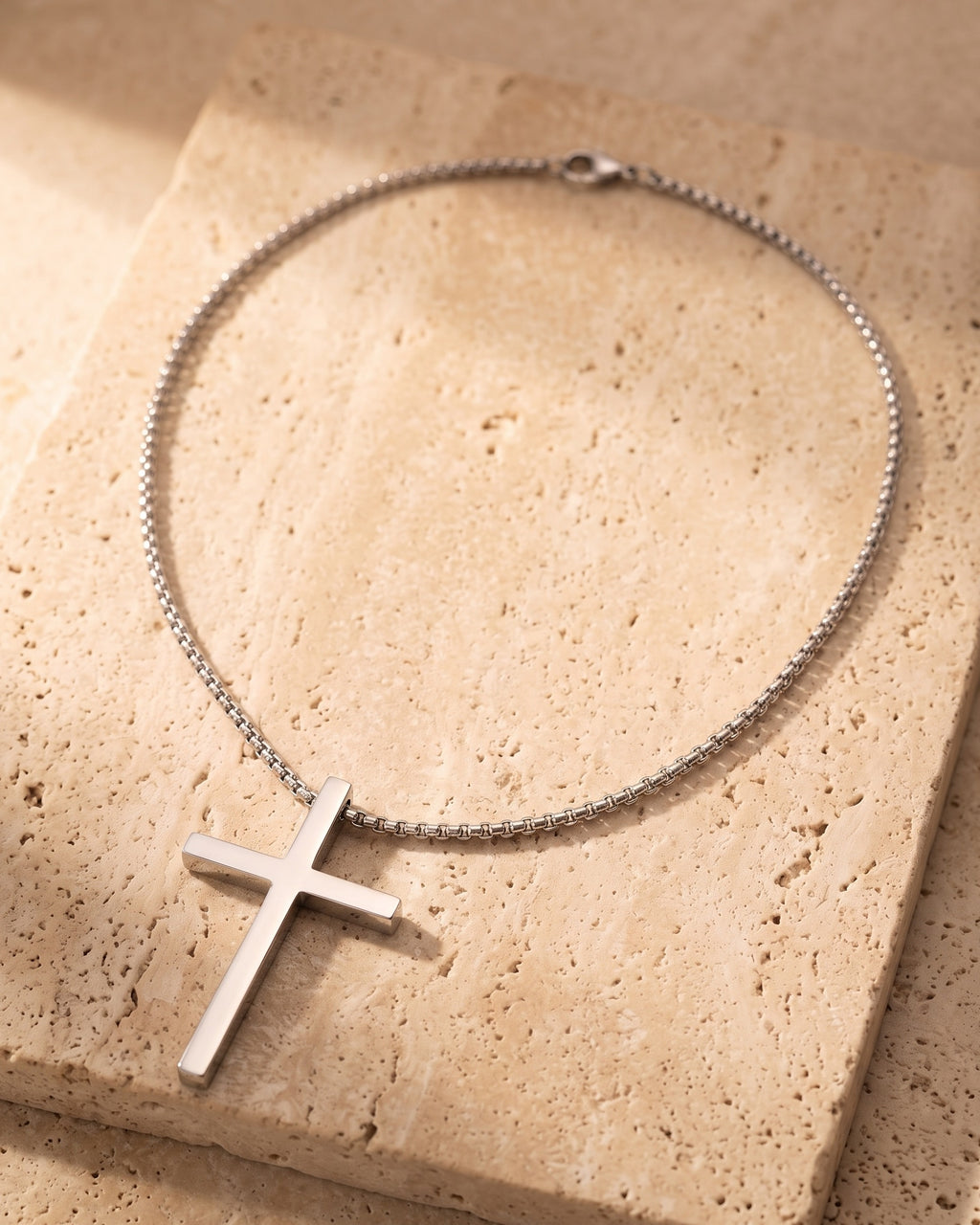 Entire Atlas minimal cross pendant necklace in silver-tone steel, lying flat on warm stone