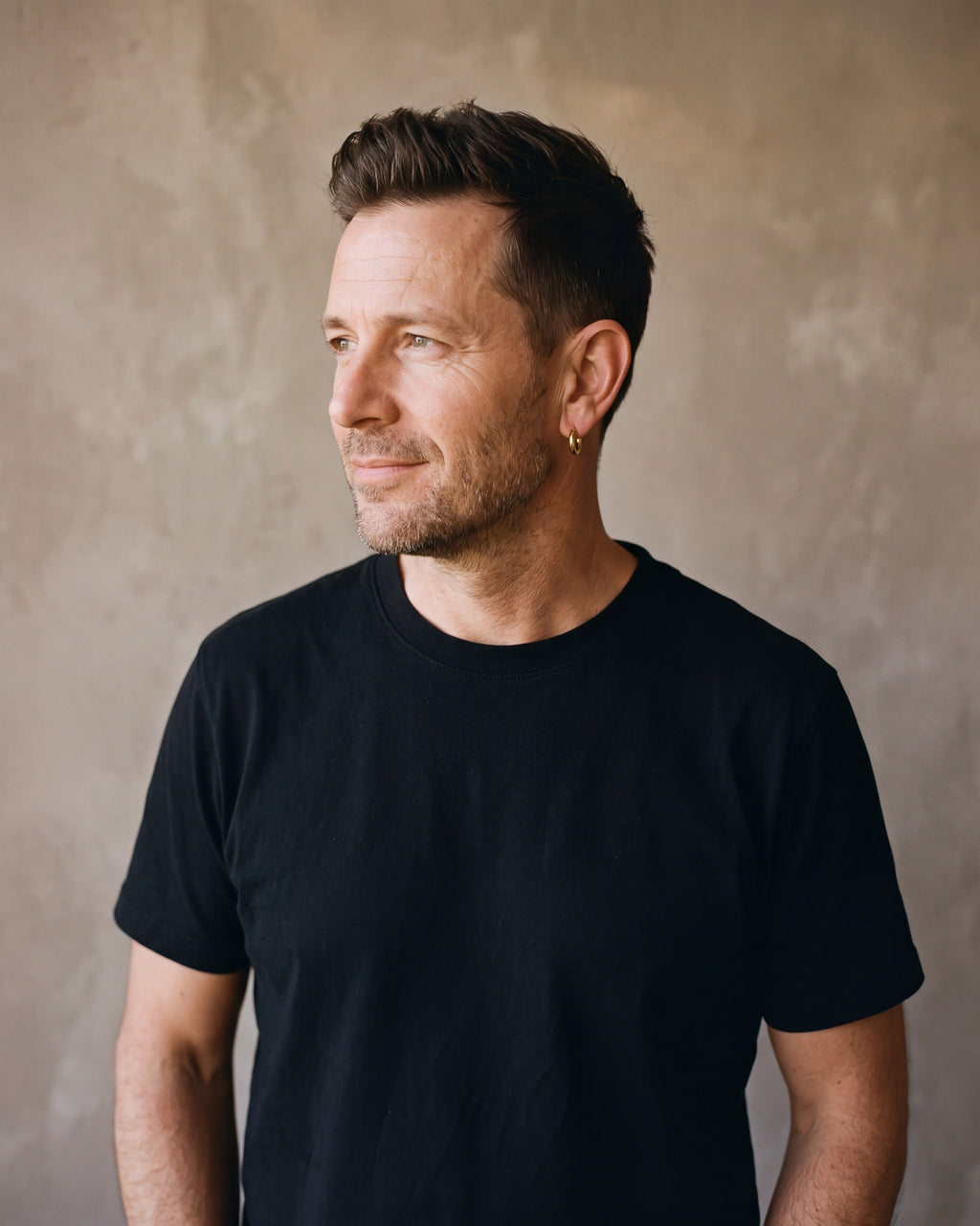 Man in a black T-shirt with his hands in his pockets wearing the gold-tone Atlas thin seamless hoop earrings
