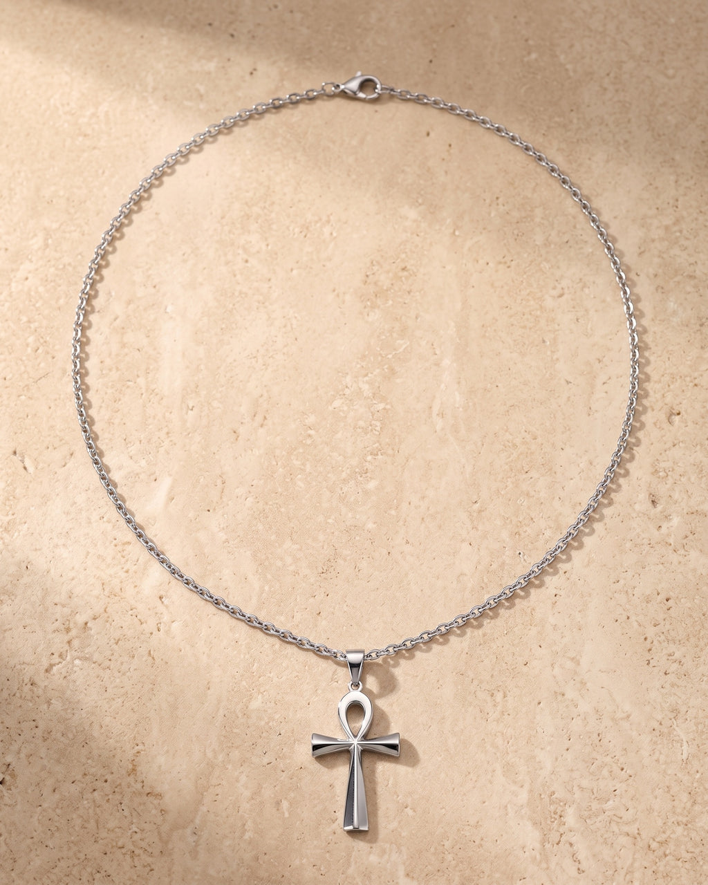 Entire Atlas ankh cross pendant necklace in silver-tone steel, lying flat on warm stone