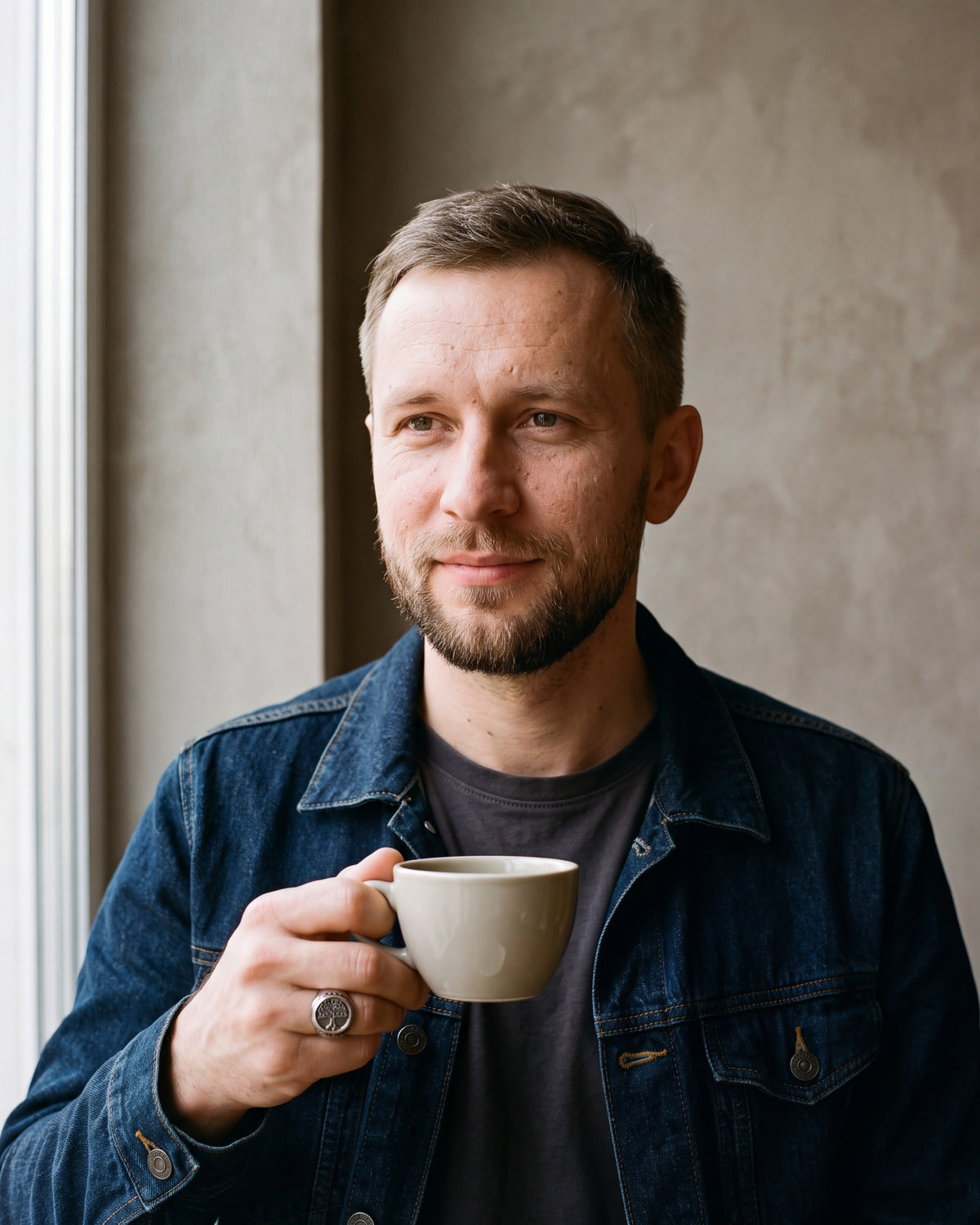 Man in a dark indigo denim jacket open over a charcoal crew-neck t-shirt by a window wearing the silver-tone Atlas tree of life signet ring