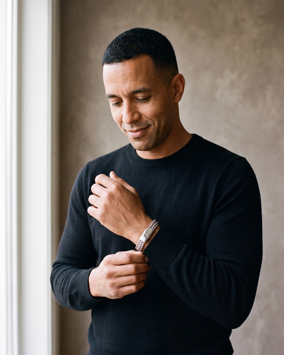 Man in a black merino crew-neck sweater by a window wearing the silver-tone Atlas twin wheat chain bracelet