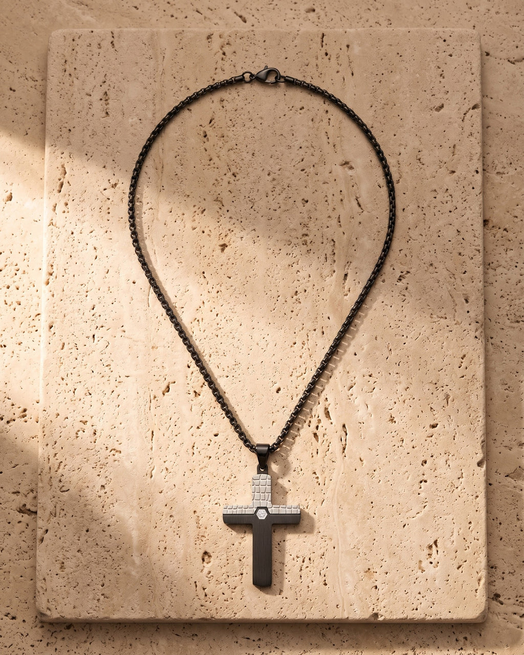 The black Atlas Two-Tone Cross Necklace with CZ laid on warm travertine stone