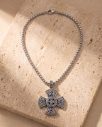 Entire Atlas Viking knotwork cross pendant necklace in silver-tone steel, lying flat on warm stone