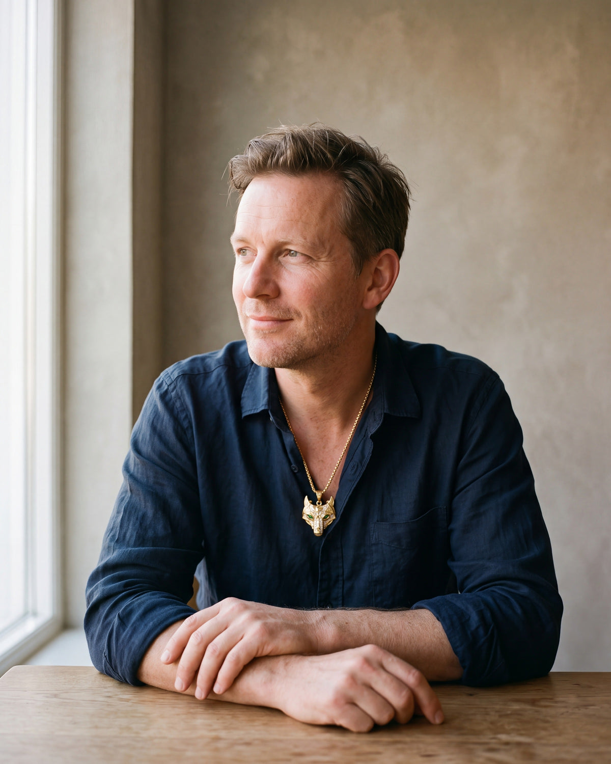 Man in a dark navy linen shirt by a window wearing the gold-tone Atlas viking wolf pendant necklace