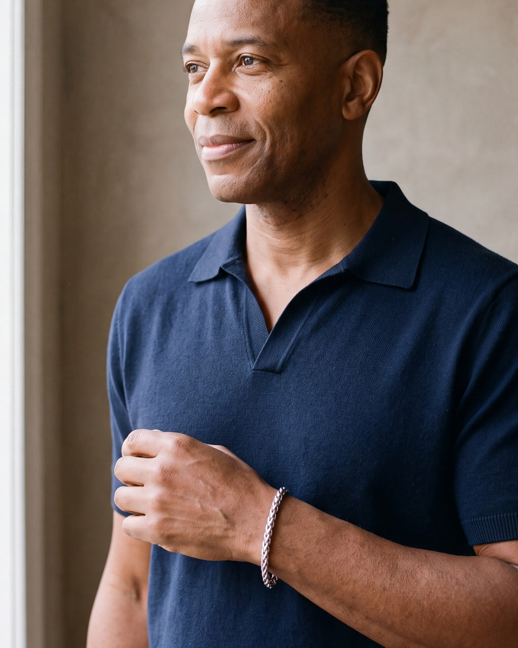 Man in a navy knit polo looking toward the window, wearing the 6 mm Atlas wheat chain bracelet