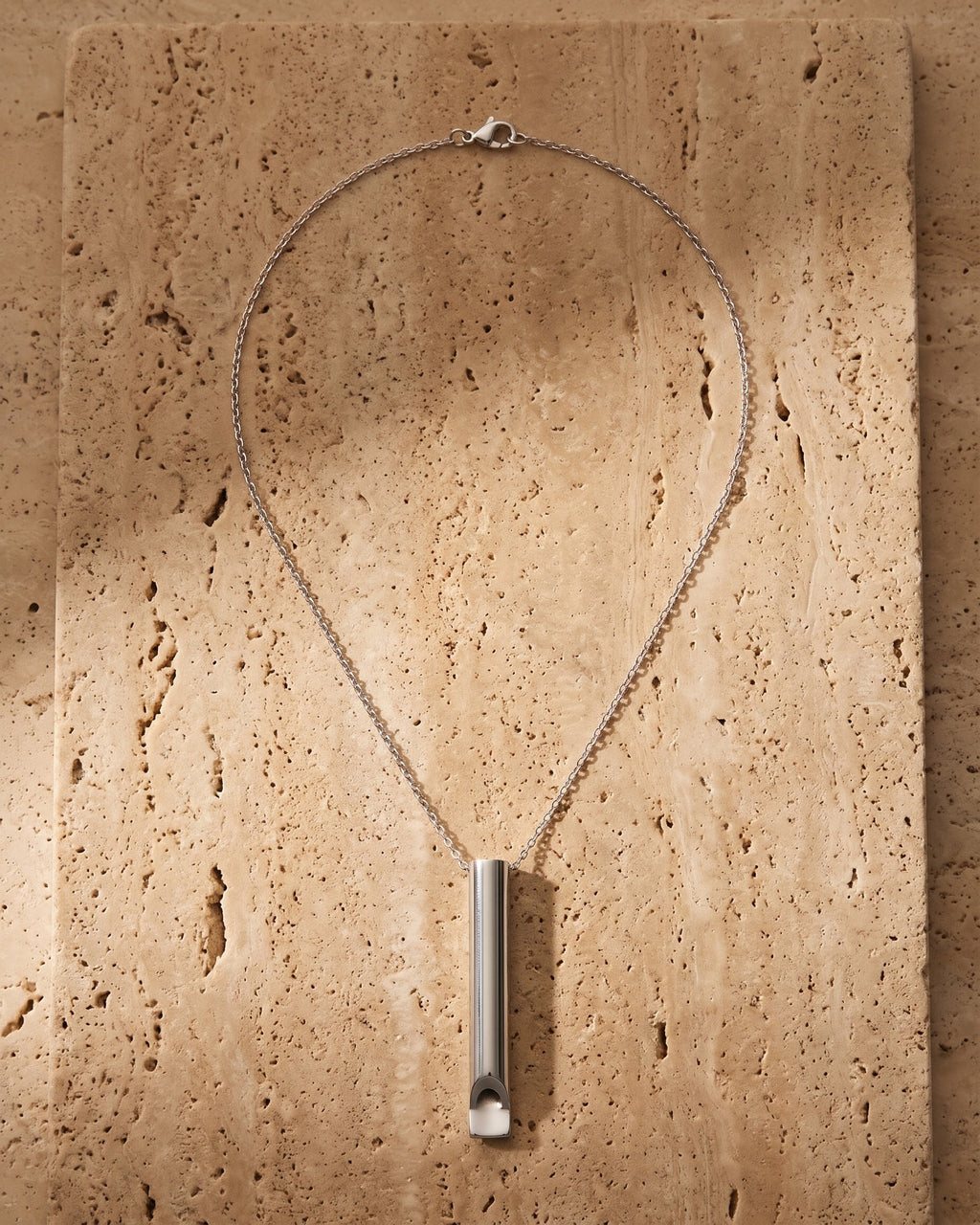The silver Atlas Whistle Pendant Necklace in Titanium Steel laid on warm travertine stone