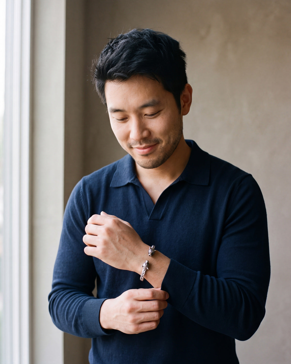 Man in a navy fine-gauge knit polo with an open collar by a window wearing the silver-tone Atlas wolf head braided cuff bracelet