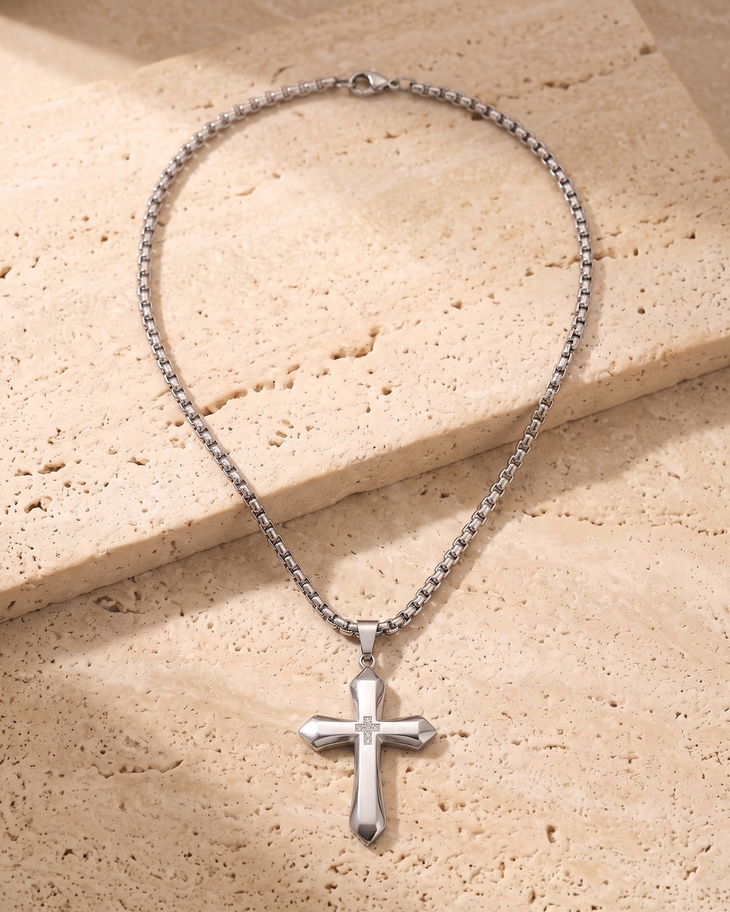Entire Atlas zircon cross pendant necklace in silver-tone steel, lying flat on warm stone