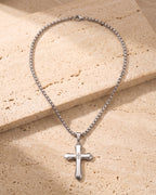 Entire Atlas zircon cross pendant necklace in silver-tone steel, lying flat on warm stone