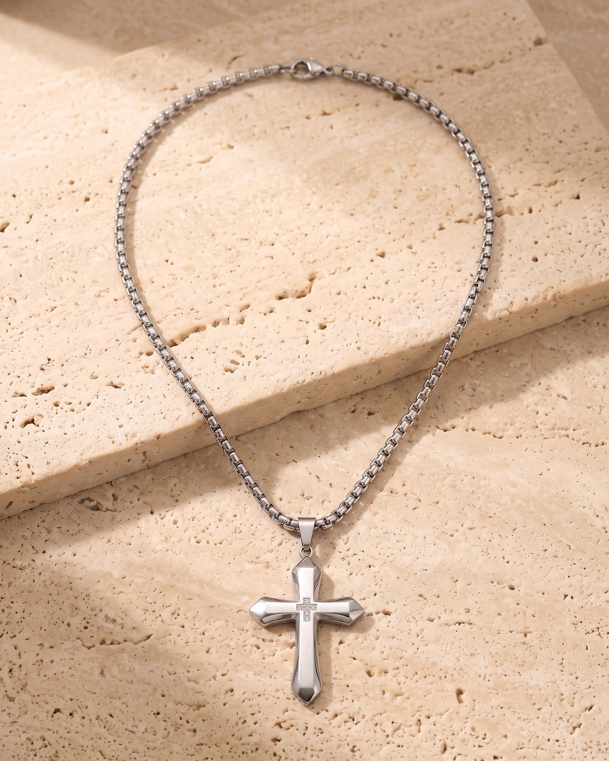 Entire Atlas zircon cross pendant necklace in silver-tone steel, lying flat on warm stone
