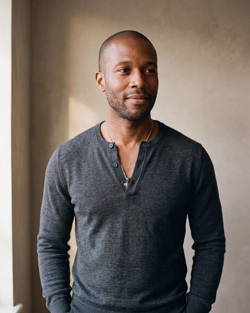 Man in a fine-gauge charcoal-grey merino knit with a henley neckline by a window wearing the silver-tone Atlas Interlocking Cross Pendant Necklace