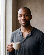 Man in a fine-gauge charcoal-grey merino knit with a henley neckline by a window wearing the silver-tone Atlas Hollow Geometric Cross Necklace