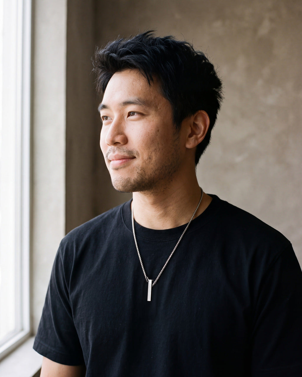 Man in a plain heavyweight black cotton crew-neck t-shirt (thick by a window wearing the silver Atlas script bar pendant necklace